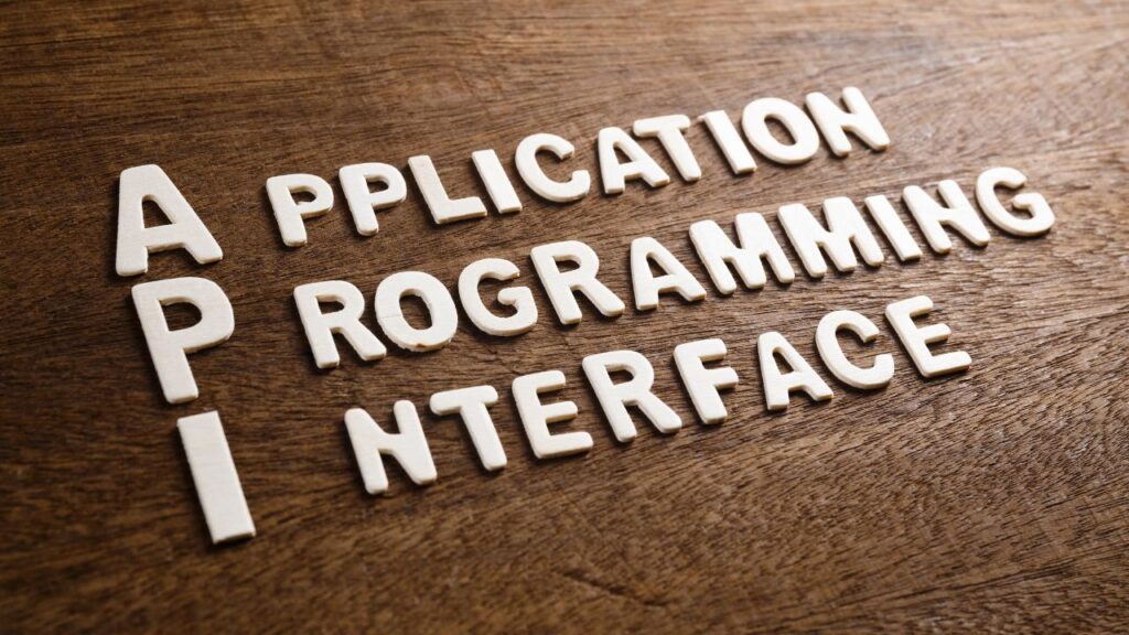 Três linhas com letras brancas formando as palavras “Application”, “Programming” e “Interface” sobre fundo de madeira, explicando o termo API.