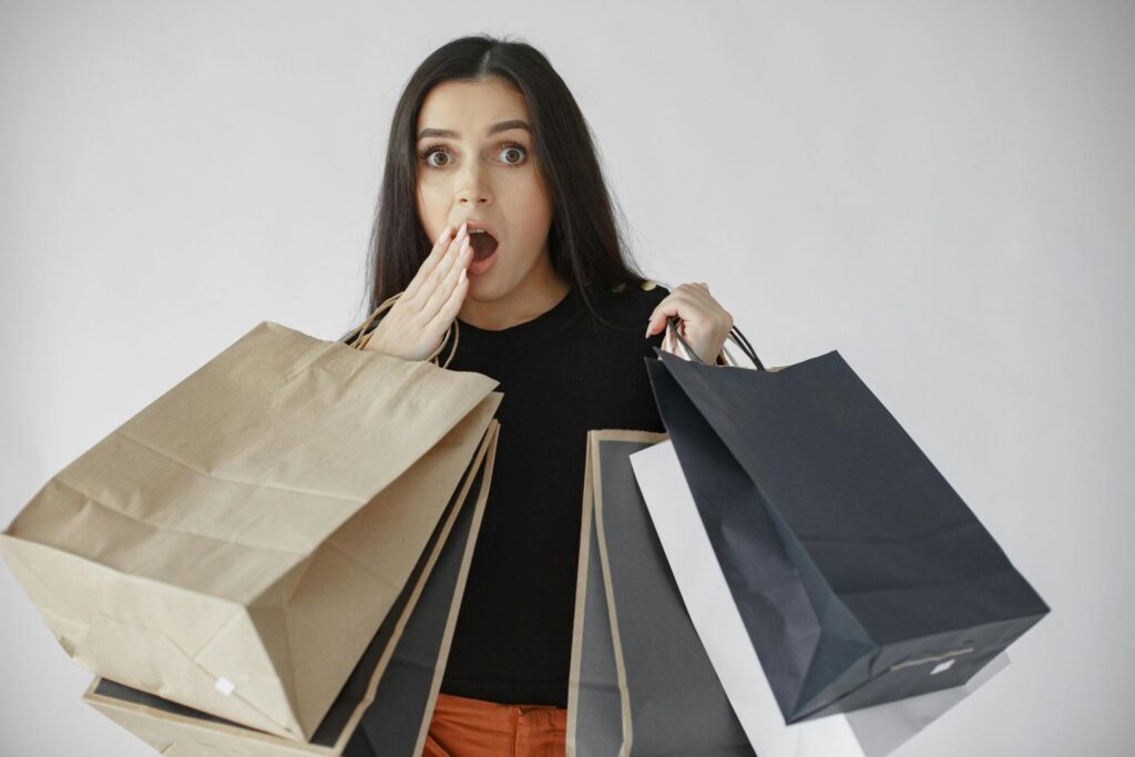 Jovem com expressão de surpresa segurando várias sacolas de compras. Palavra-chave: jovem consumidor