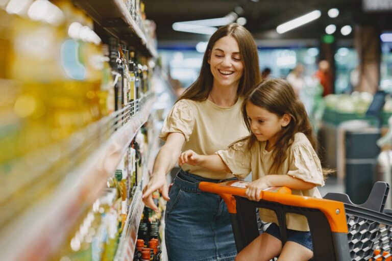 Mãe sorridente apontando prateleira de mercado enquanto filha observa atenta, simbolizando influência geracional no consumo. Palavra-chave: jovem consumidor